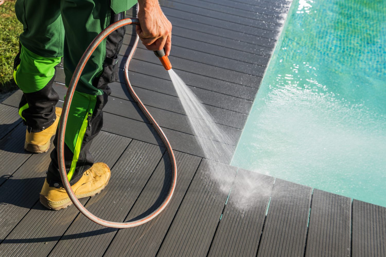 Garden Maintenance Worker Washing the Timber Decking Around Residential Swimming Pool with a Hose.